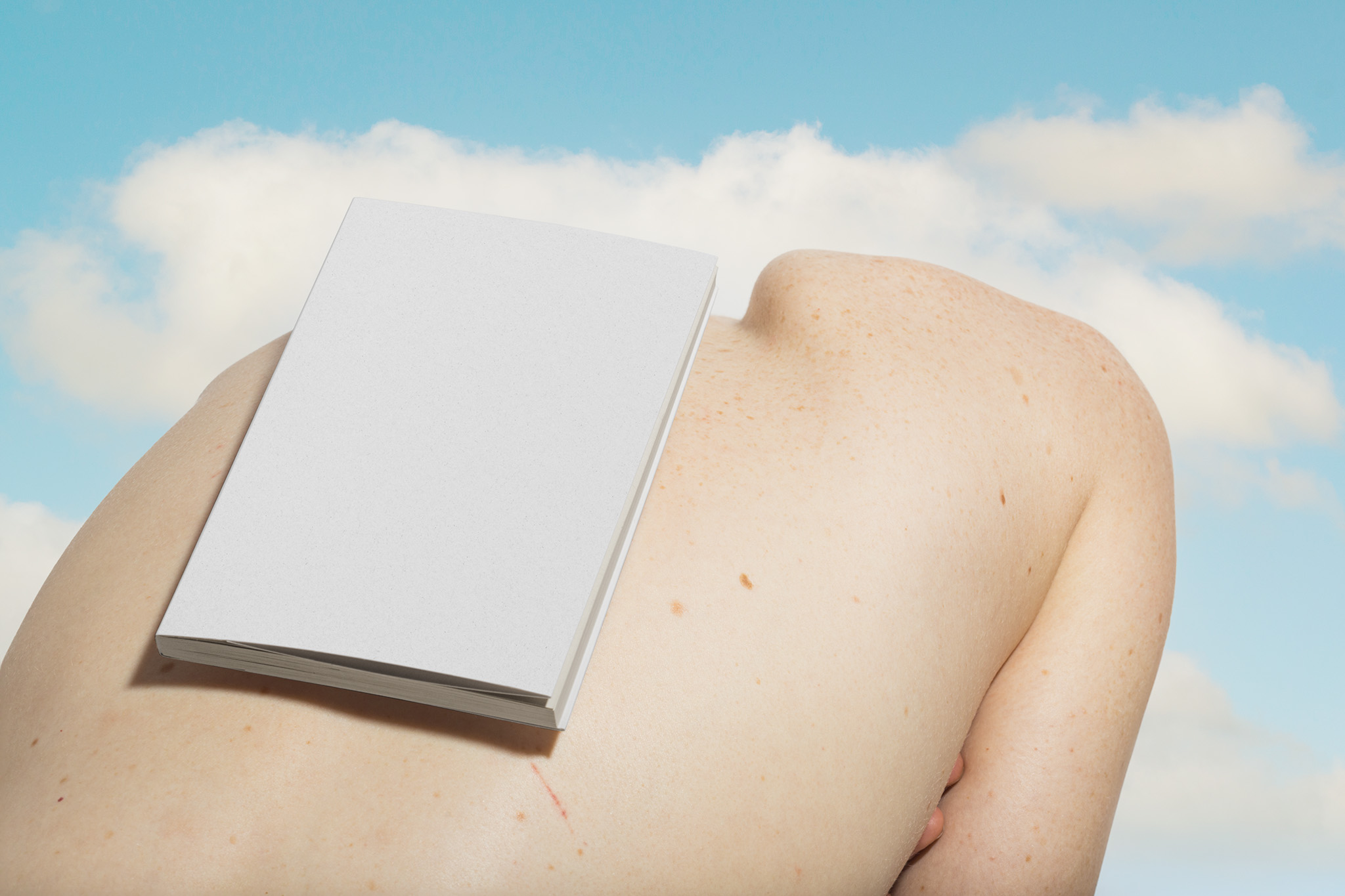 Slouched back bodyscape featuring an A5 book softcover mockup, set against a cloudy blue sky, empty mockup.