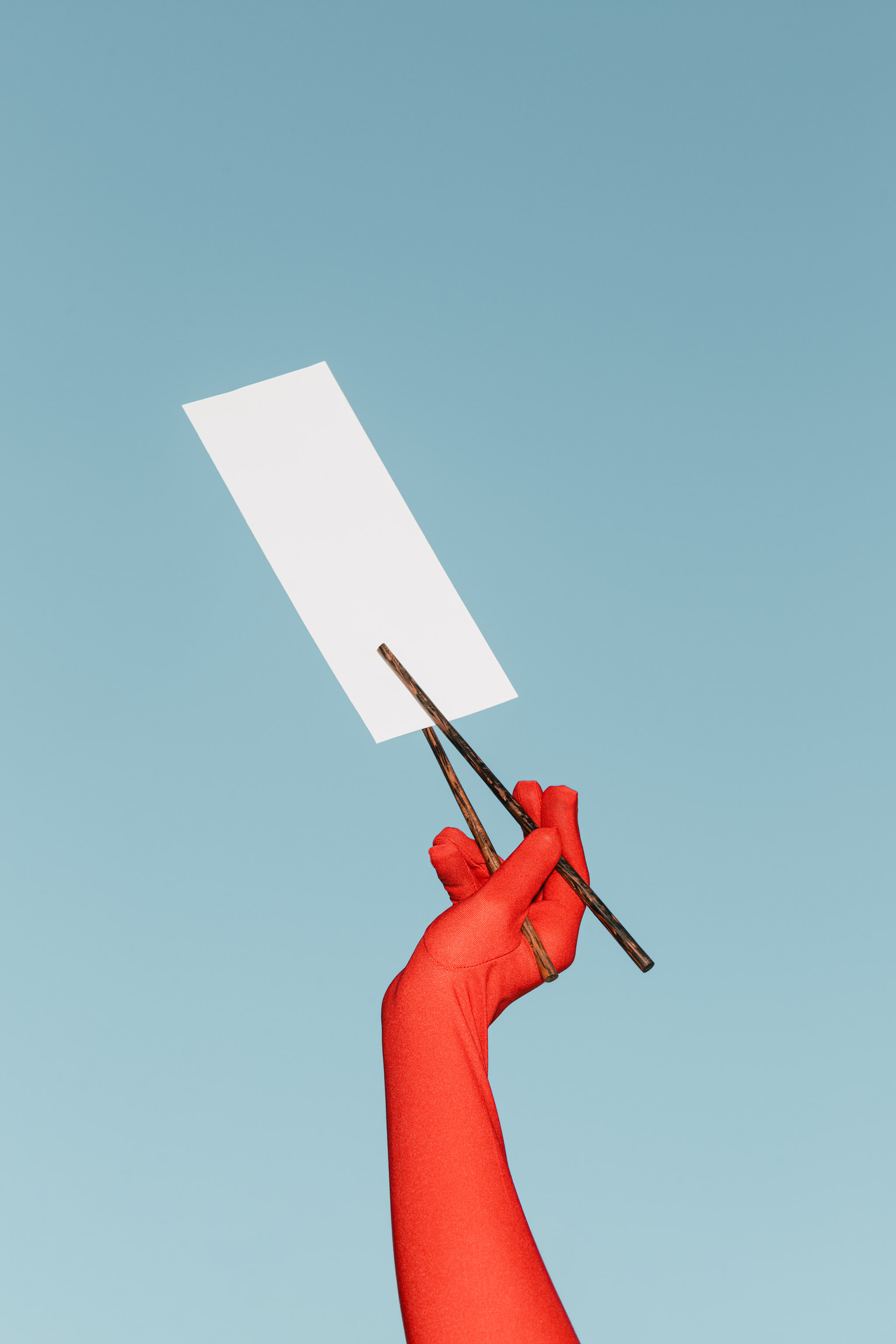 Photo of a red gloved hand, set against a light pastel blue sky, holding up a vertical paper menu with black typography with dark wooden chopsticks, empty mockup.