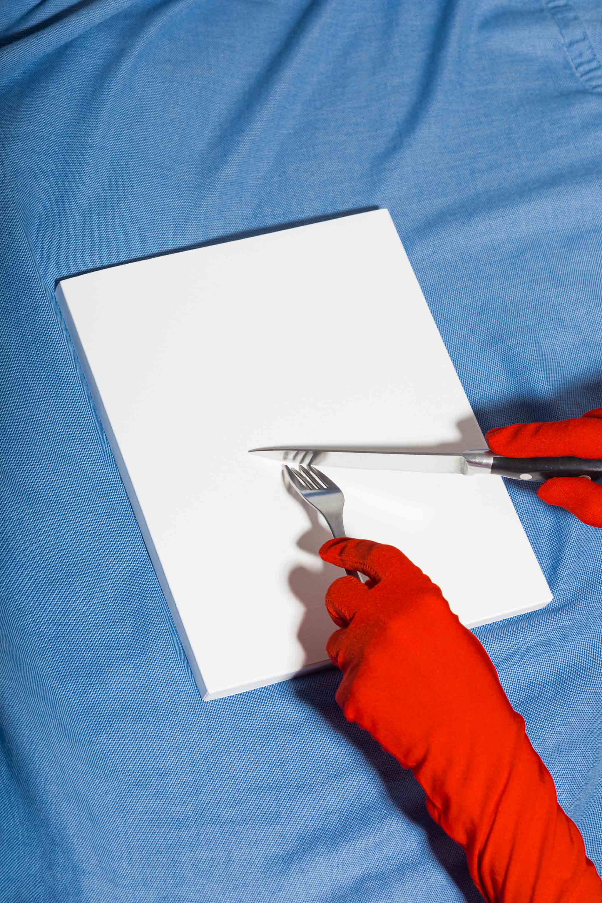 Two gloved hands holding utensils over a book cover mockup set against blue fabric, empty mockup.