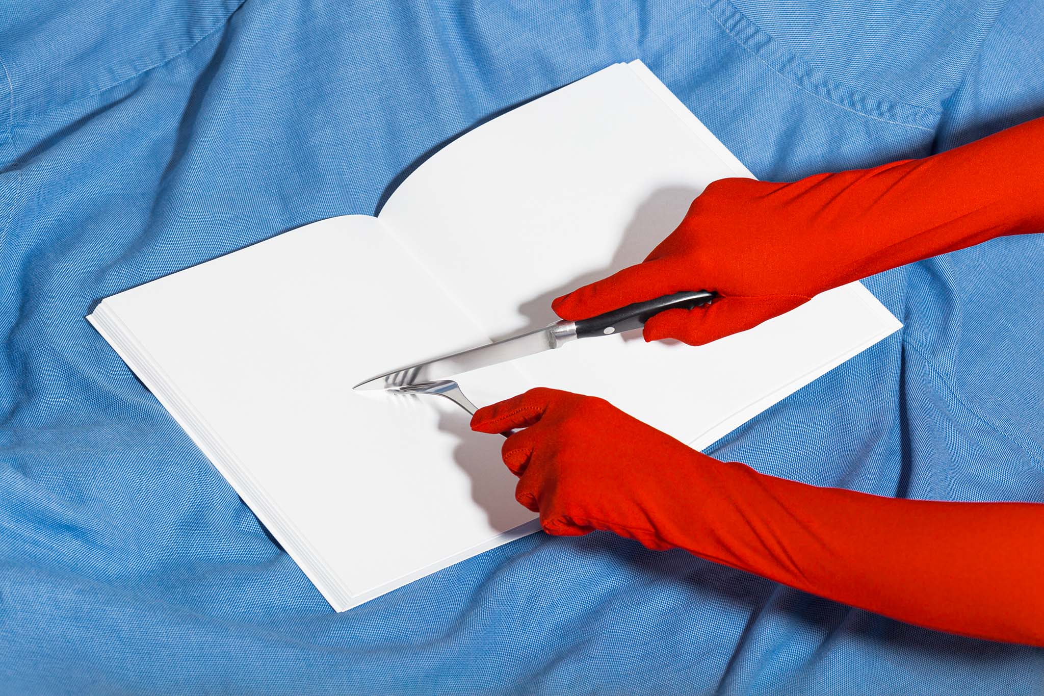 Two gloved hands holding utensils over an open book mockup set against light blue fabric, empty mockup.
