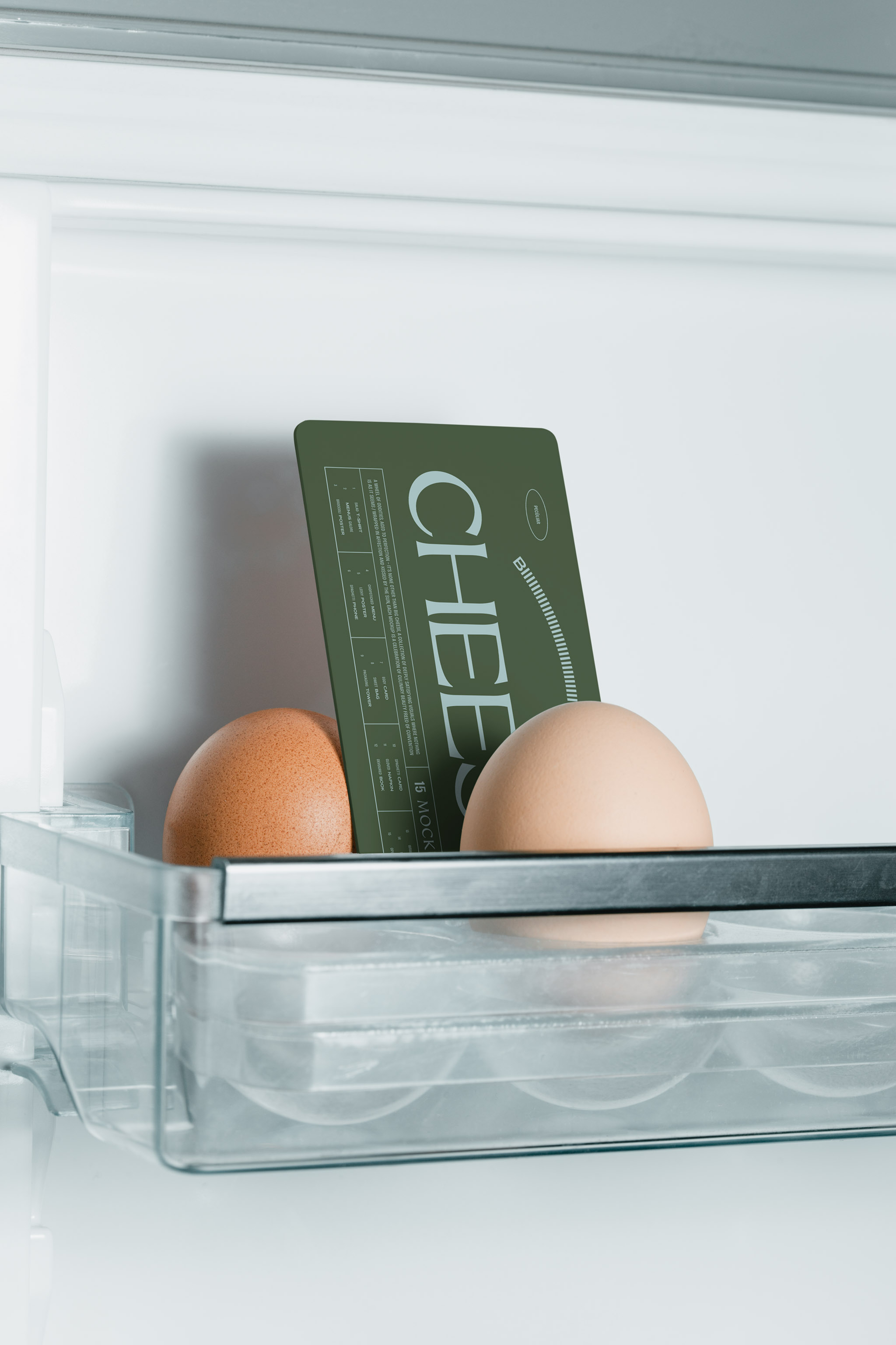 Close-up photo of two eggs in a minimalist white fridge among which a vertical american business card mockup with dark typography is placed, in use example alternative colors.
