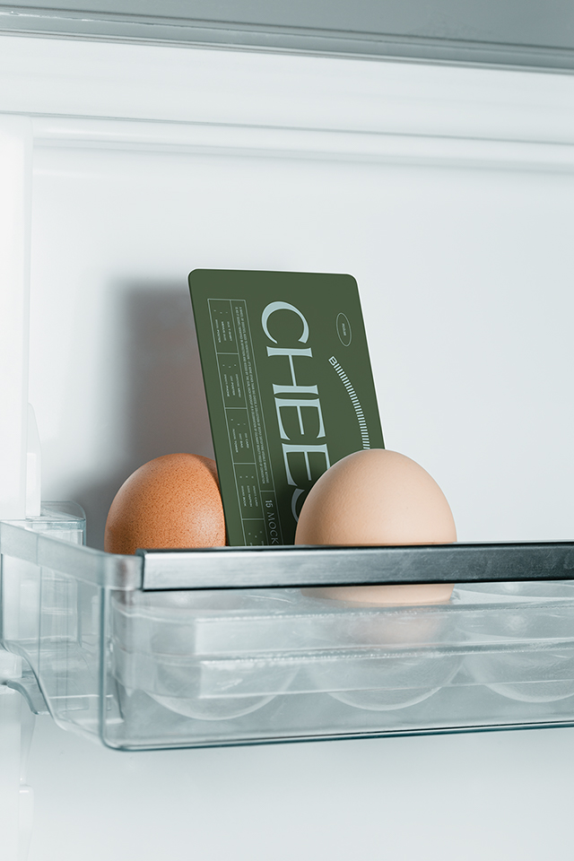 Close-up photo of two eggs in a minimalist white fridge among which a vertical american business card mockup with dark typography is placed, empty mockup.