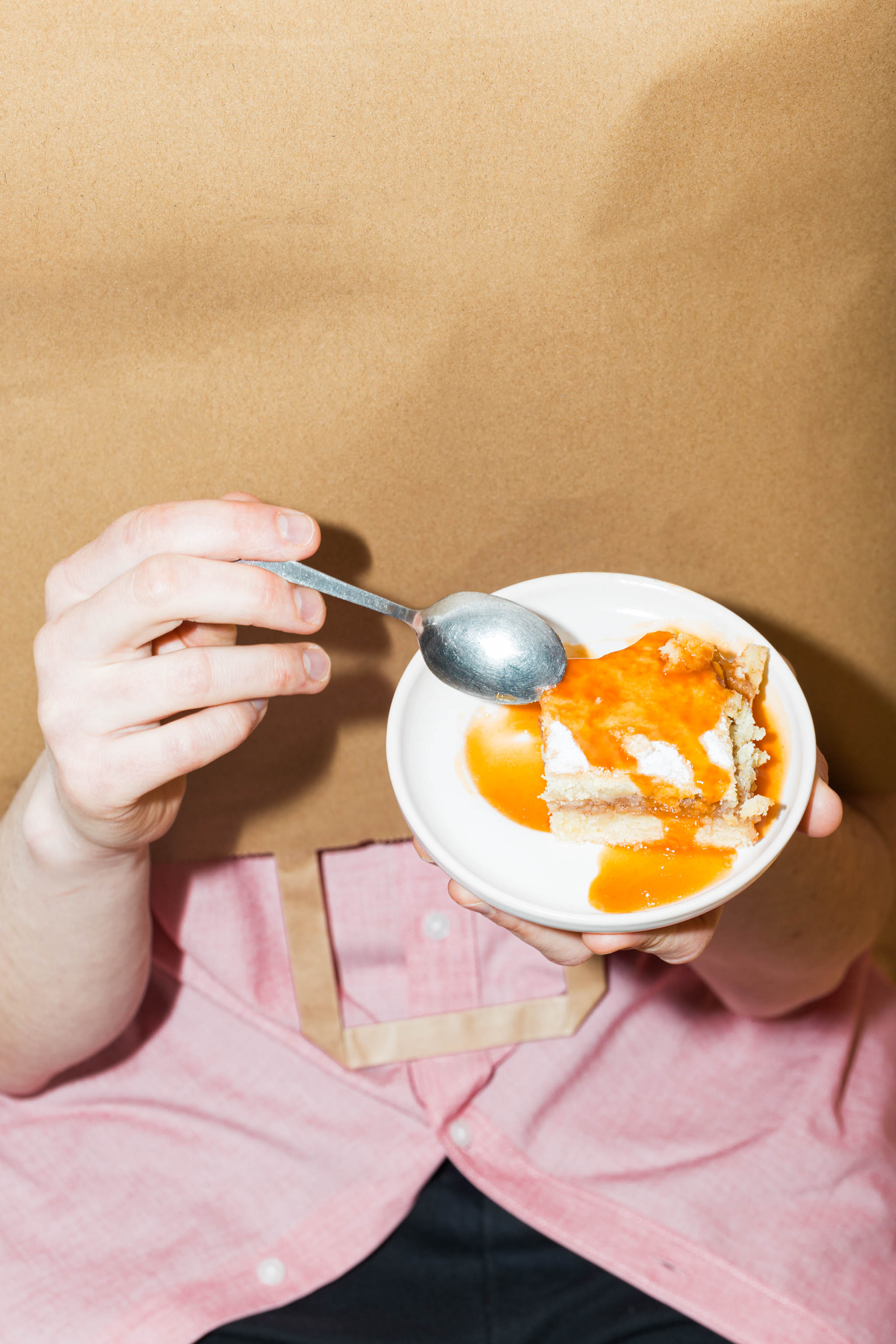 Close-up photo of paper bag mockup turned upside-down over a person's head while they're holding up a sweet orange dessert in a pink dress shirt and black pants, empty mockup.