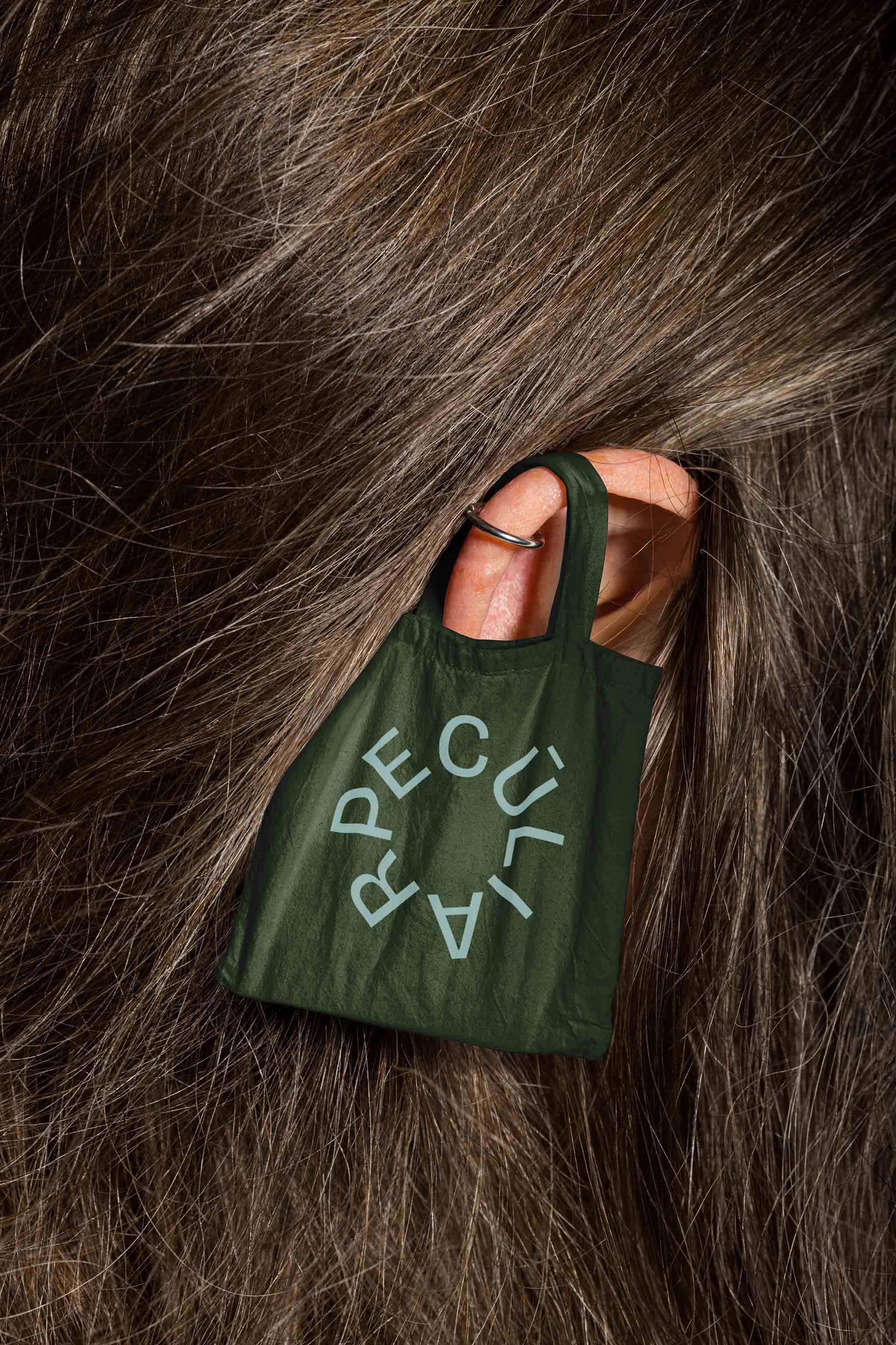 Canvas tote bag mockup hung on a person's ear amid dark wavy hair, in use example alternative colors.