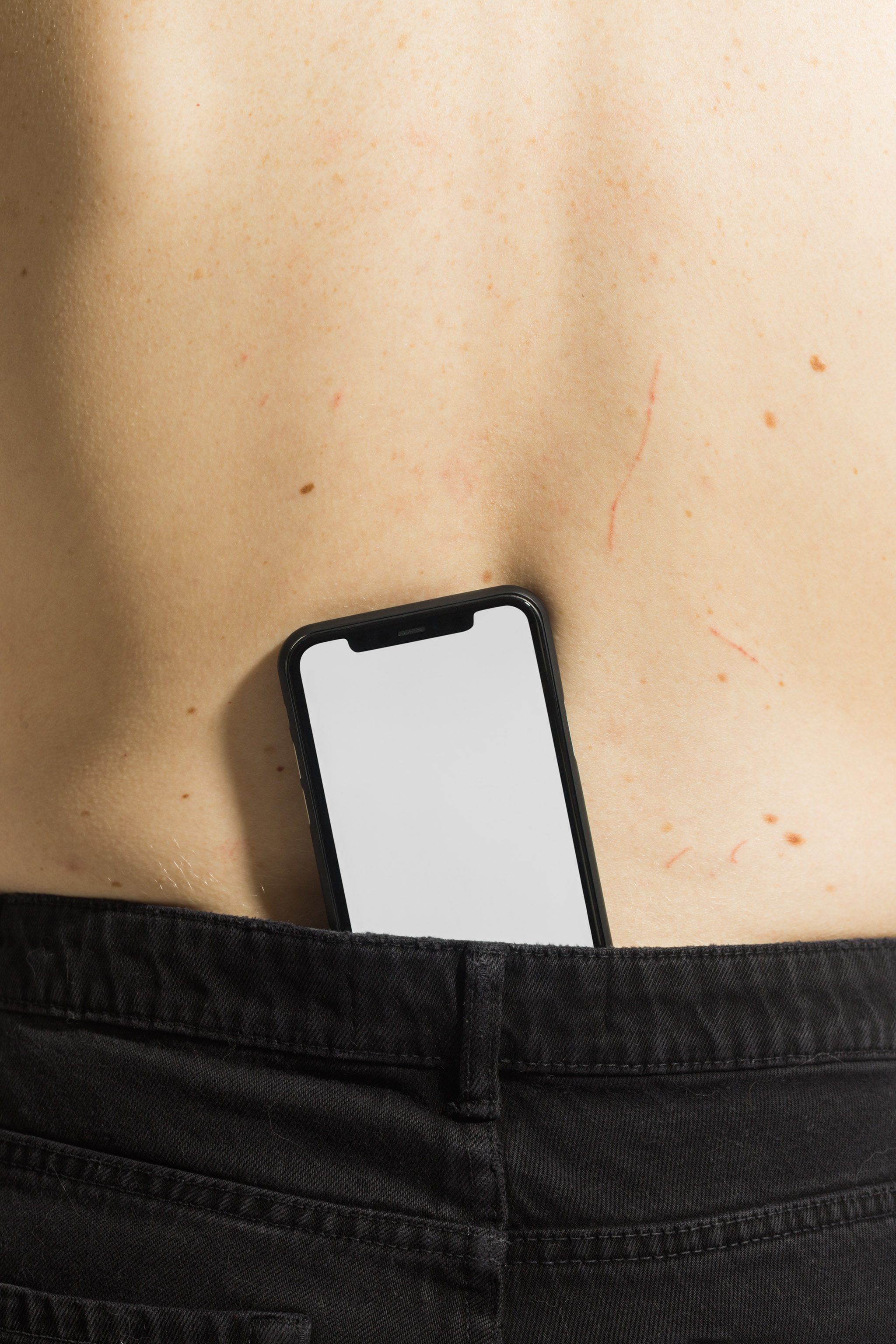 Close-up mockup of a black iPhone on bare skin tucked behind jeans, empty mockup.